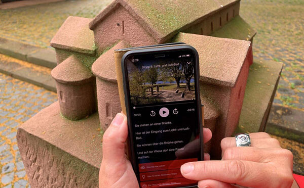Foto von einem Handy mit der App vom Pilgerweg. Hinter dem Handy: ein Modell einer Kirche aus Stein.