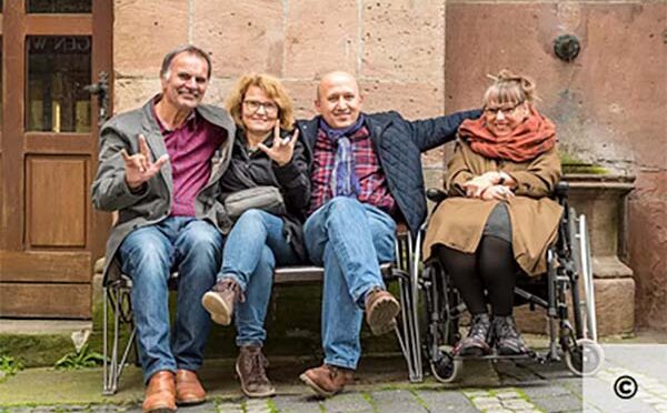 Vier lachende Menschen sitzen vor einem alten Haus aus Stein. Eine Dame im Rollstuhl, ein Pärchen macht ein Zeichen aus der Gebärdensprache
