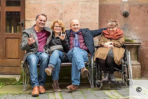 Vier lachende Menschen sitzen vor einem alten Haus aus Stein. Eine Dame im Rollstuhl, ein Pärchen macht ein Zeichen aus der Gebärdensprache