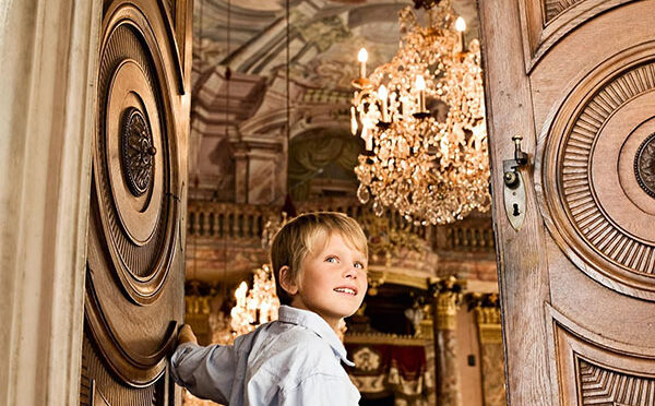 Ein Junge macht eine Tür auf zu einem festlichen Saal mit edlem Kronleuchter.